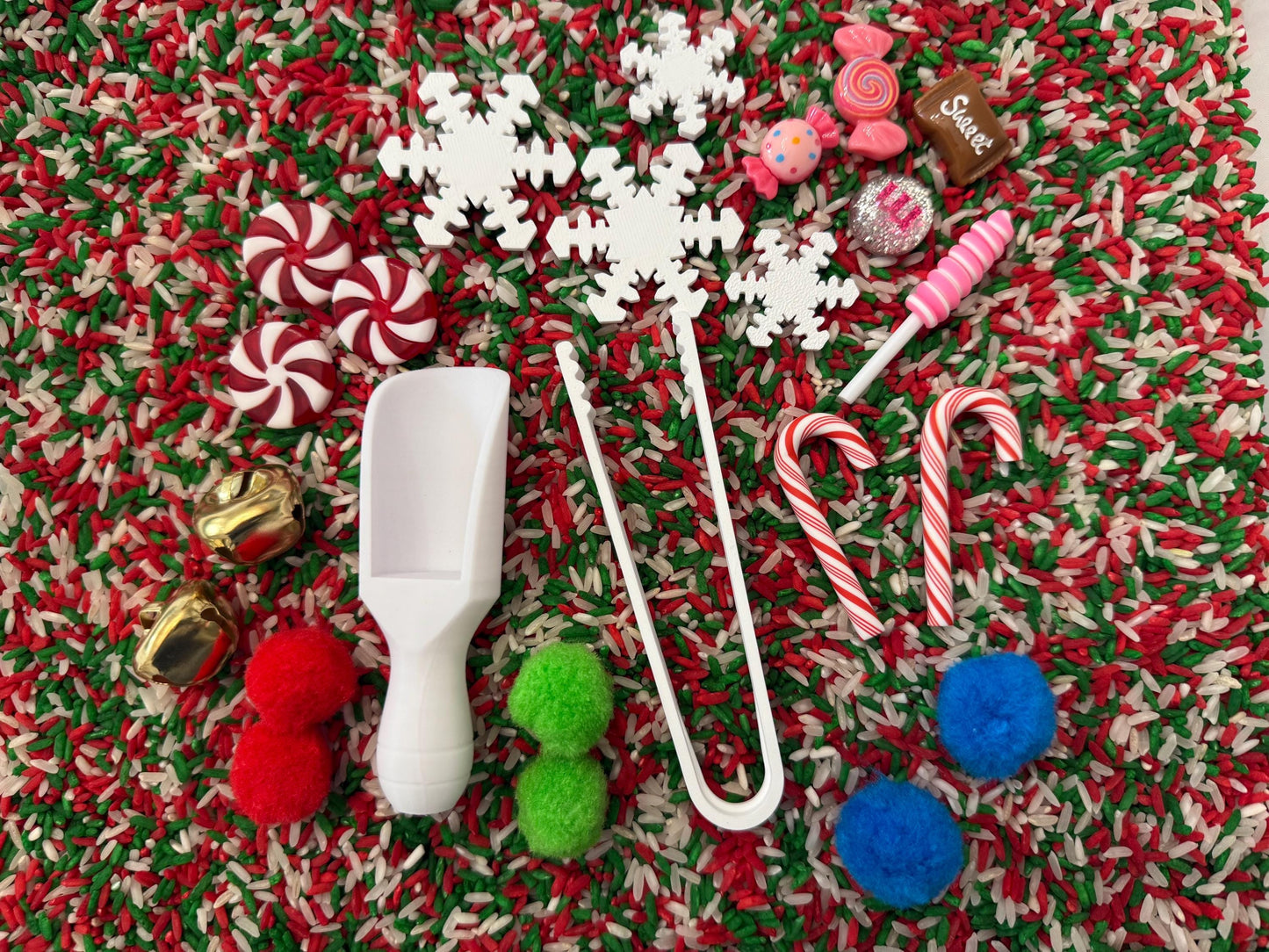 May include: A festive sensory bin with a red, green, and white rice base. Includes white snowflakes, candy, pom-poms, a scoop, tongs, and candy canes. Gold bells and a candy-shaped object are also present.