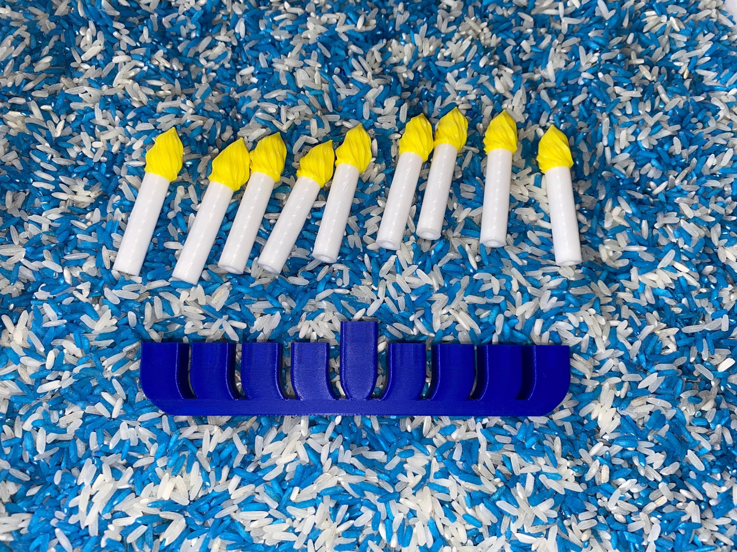 May include: A blue plastic menorah with eight yellow candles on a bed of blue and white rice.