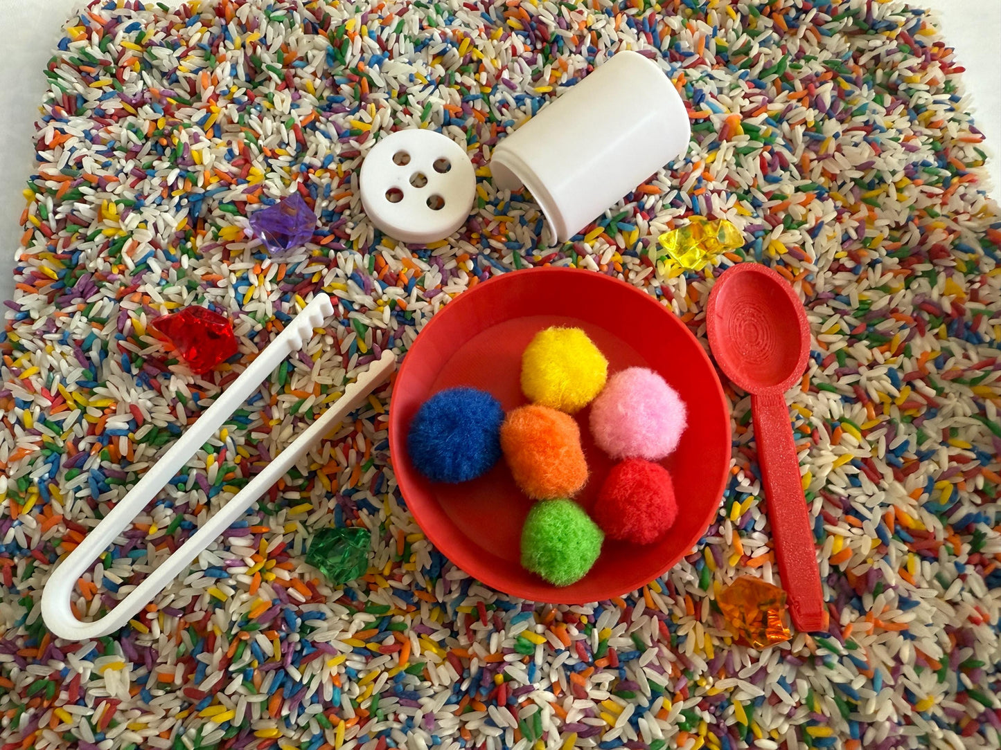 May include: A red bowl filled with colorful pom-poms sits on a tray of rainbow rice. A white plastic spoon, tongs, and a small white container with holes are also on the tray.