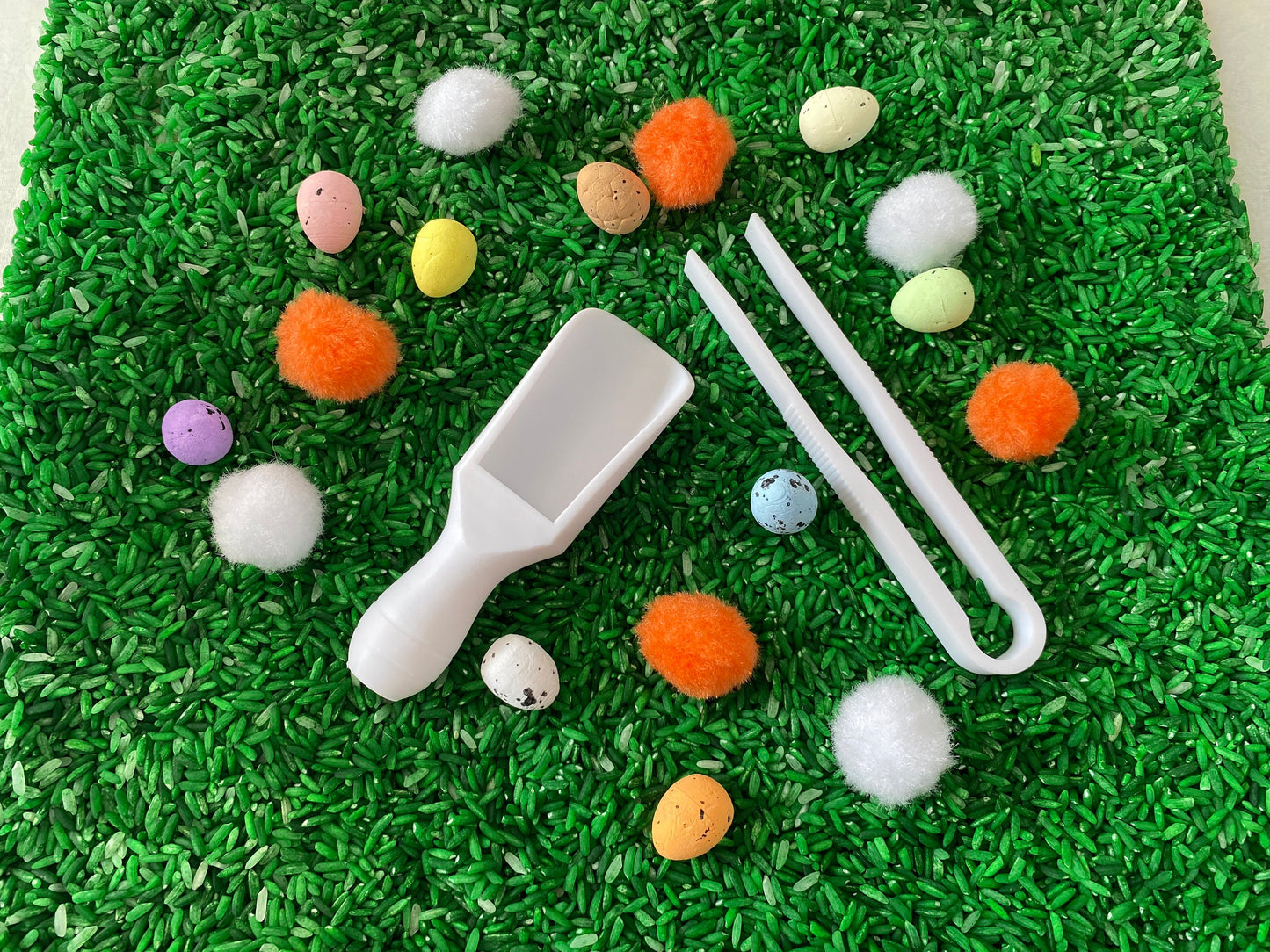 May include: A white plastic scoop and tweezers are on a bed of green rice. There are also several small colorful eggs and pom-poms scattered around.
