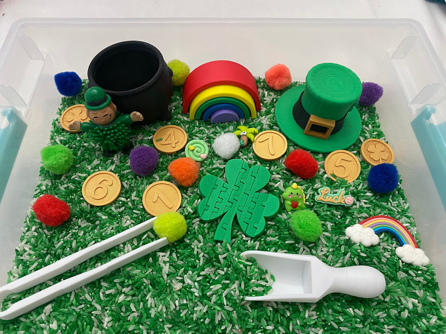 May include: A green sensory bin filled with green and white rice, green pom-poms, and gold coins. There is a green leprechaun figurine, a green hat, a rainbow, a green four-leaf clover, and a white scoop.