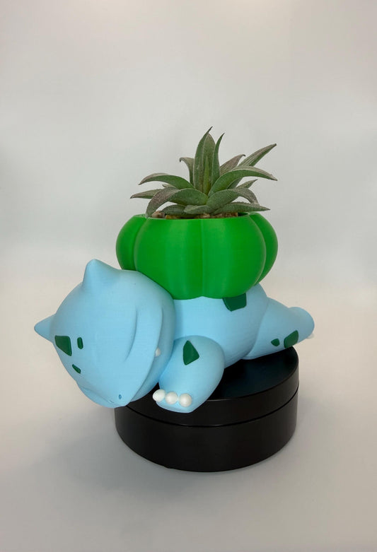 May include: A light blue Bulbasaur planter with a green bulb on its back, holding a small succulent plant. The planter is resting on a black base. The Bulbasaur has green spots and white toes.