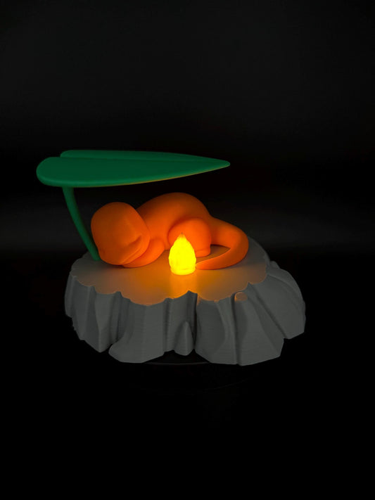 May include: An orange figurine of a creature resting under a green leaf, atop a gray, rocky base. A yellow light emanates from the center, creating a warm glow. The design is a stylized representation of a popular character.