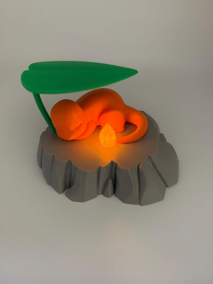 May include: An orange figurine of a creature resting on a gray, rock-like base, with a green leaf overhead. A small, glowing orange light sits near the creature. The figurine is a stylized representation of a popular character.