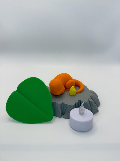 May include: An orange creature figurine resting on a gray rock formation, with a yellow flame and a green leaf. A white, battery-operated candle is also present. The objects are arranged on a white surface.