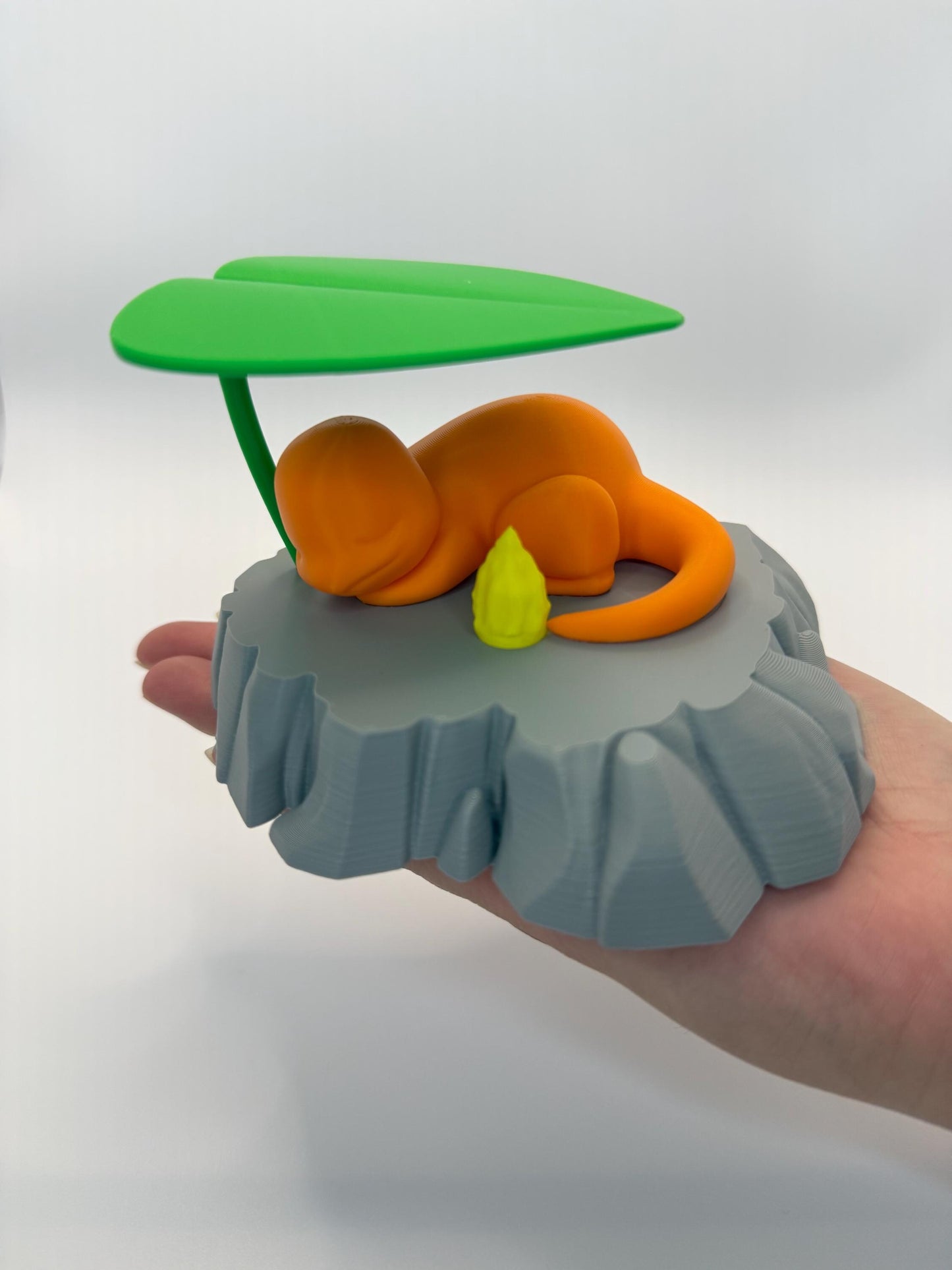 May include: An orange figurine of a Charmander character resting on a gray rock formation, with a green leaf overhead. A small yellow flame is next to the character. The figurine is a 3D-printed item.