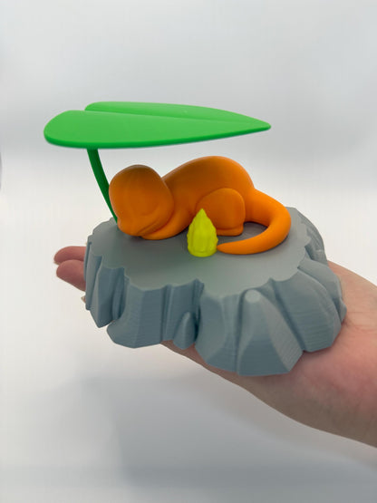 May include: An orange figurine of a Charmander character resting on a gray rock formation, with a green leaf overhead. A small yellow flame is next to the character. The figurine is a 3D-printed item.