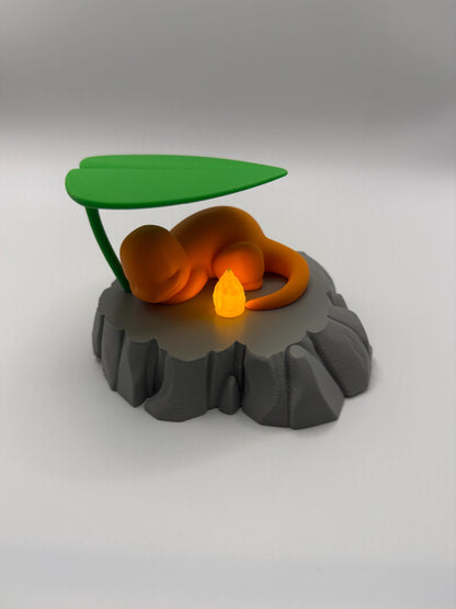May include: A 3D-printed orange creature figurine resting on a gray, rocky base, under a green leaf. A small, glowing orange light sits near the creature. The figurine is a stylized representation of a popular character.