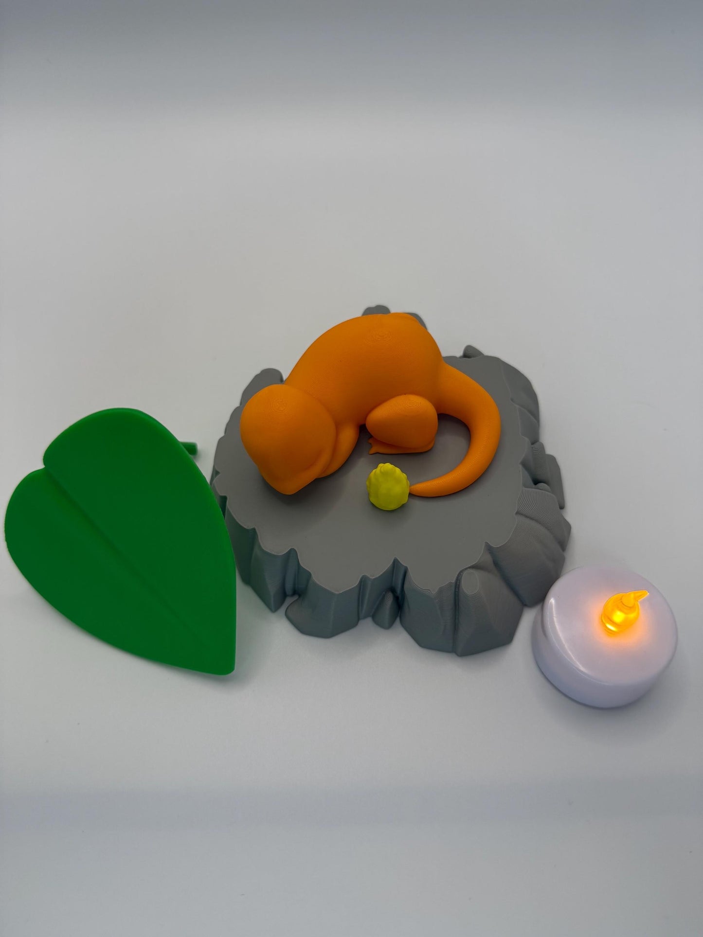May include: An orange cartoon-style figure rests on a gray, rock-shaped base, with a small yellow object nearby. A green leaf and a small, white candle with a yellow light are also present. The scene is set against a white background.