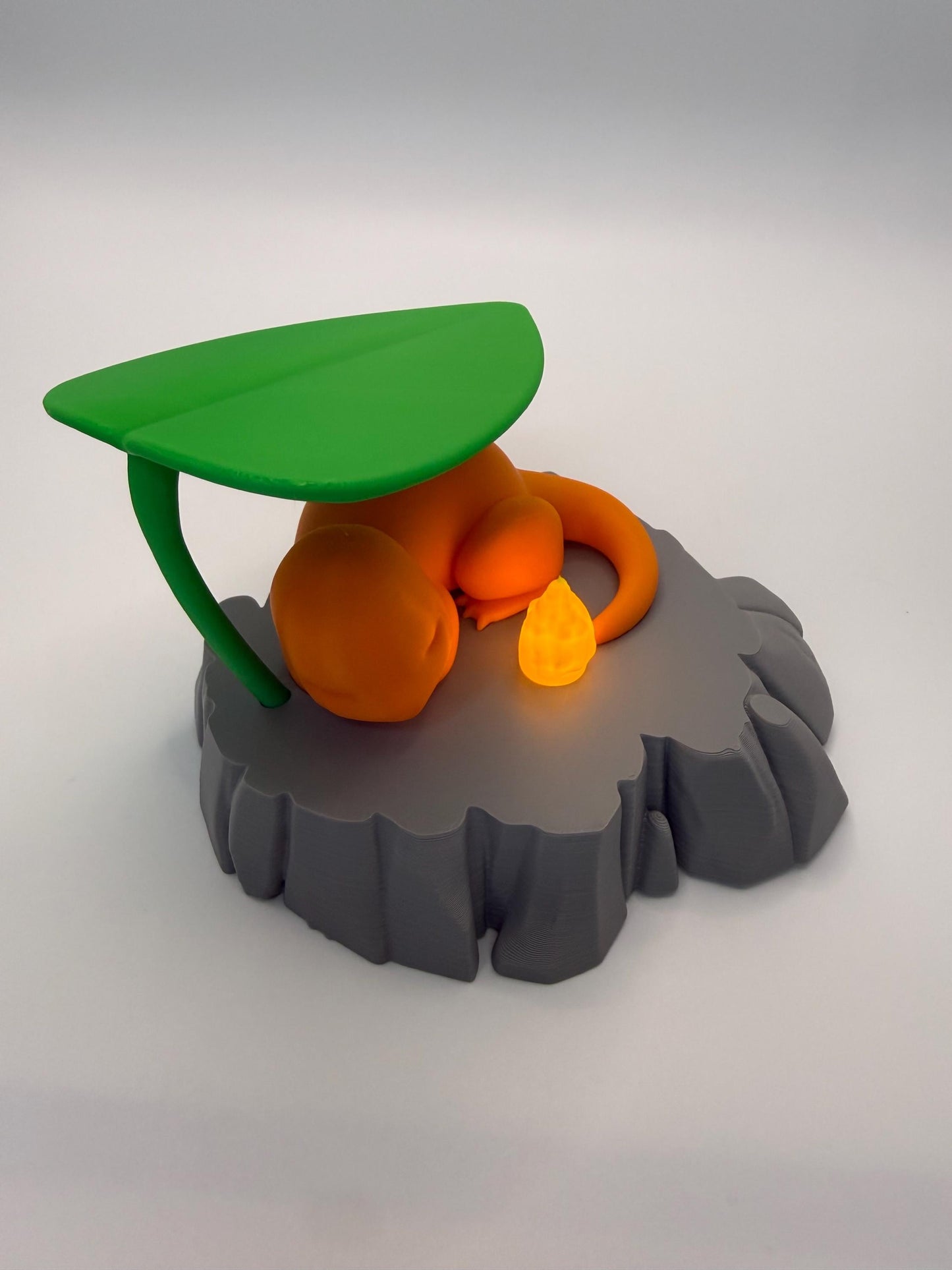 May include: A 3D-printed orange figure of a Charmander resting on a gray rock-like base, with a green leaf overhead. A small, glowing orange flame sits nearby. The design is a stylized representation of a popular character.