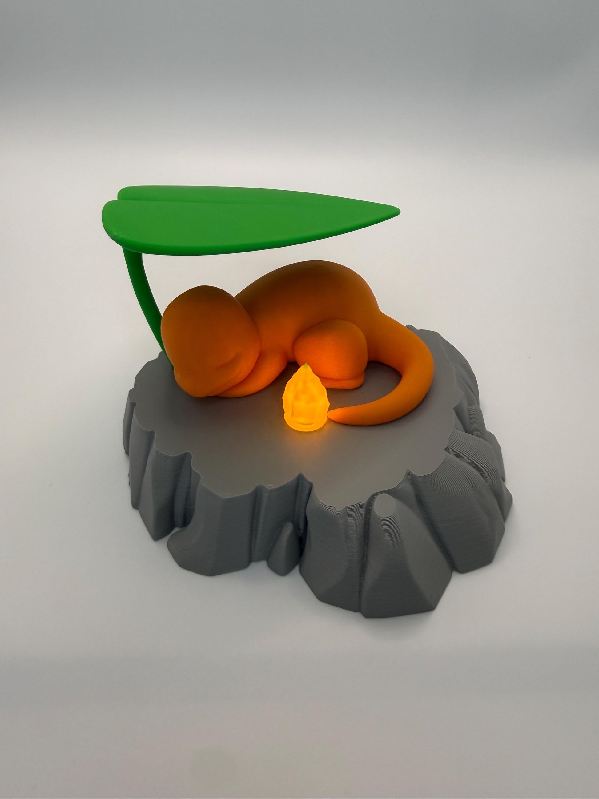 May include: An orange figurine of a sleeping creature with a flame at the end of its tail, resting on a gray rock-like base. A green leaf provides shade. The flame glows with a warm, yellow light, creating a whimsical, fantasy-themed decorative piece.