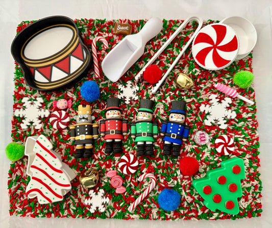 May include: A festive Christmas-themed sensory bin with a variety of toys and decorations. Includes toy soldiers, a drum, candy canes, snowflakes, and a green Christmas tree. The background is a mix of red, green, and white sprinkles.