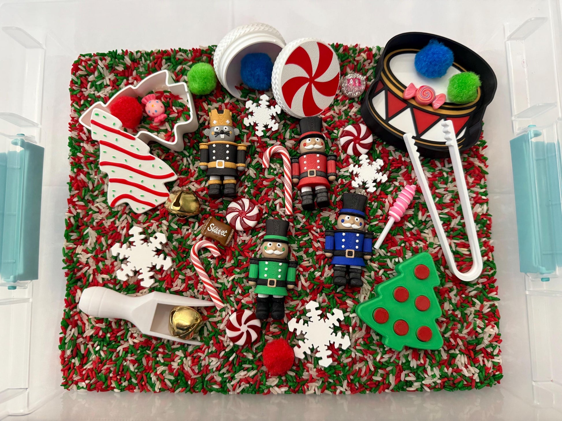May include: A Christmas-themed sensory bin filled with red, green, and white rice. Includes toy nutcrackers, candy canes, snowflakes, a drum, and a Christmas tree-shaped cookie cutter. A fun and festive activity for children.