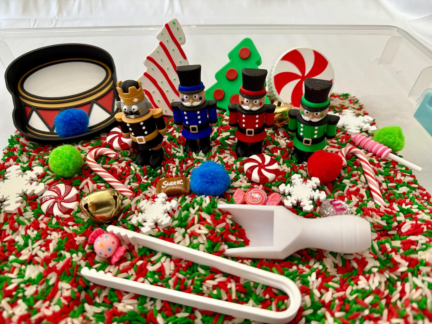 May include: A festive sensory bin with a Christmas theme. It features toy nutcrackers, a drum, candy canes, and decorative elements like snowflakes and pom-poms. The base is a mix of red, green, and white rice. A white scoop and tongs are also included.