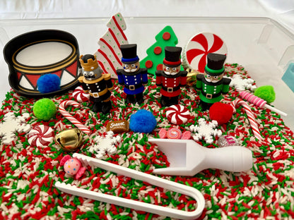 May include: A festive sensory bin with a Christmas theme. It features toy nutcrackers, a drum, candy canes, and decorative elements like snowflakes and pom-poms. The base is a mix of red, green, and white rice. A white scoop and tongs are also included.