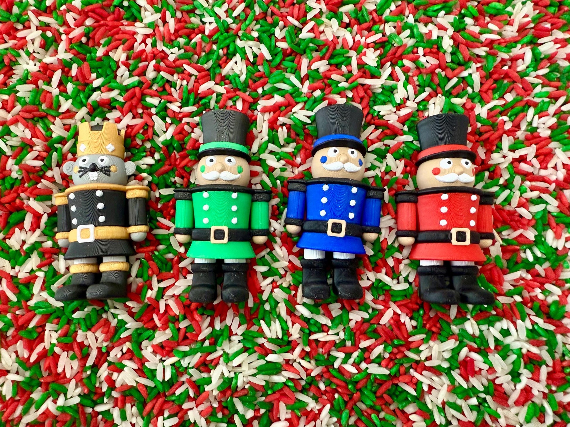 May include: Four colorful toy nutcracker figures in black, green, blue, and red uniforms with black boots, set against a background of red, green, and white sprinkles. Each nutcracker has a different hat and facial features.