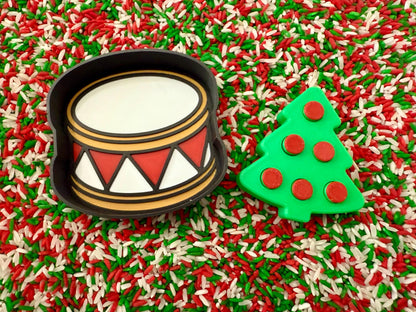 May include: A black drum-shaped cookie cutter with a white drum face, red and white triangles, and gold trim. A green Christmas tree cookie cutter with red circle ornaments is next to the drum. The background is a mix of red, green, and white sprinkles.