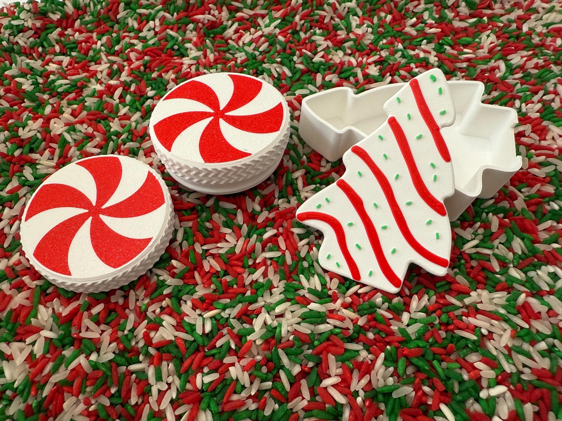 May include: Two white and red peppermint-style circles and a white Christmas tree-shaped cookie cutter with red stripes and green sprinkles are displayed on a bed of red, green, and white rice. The items are festive and holiday-themed.