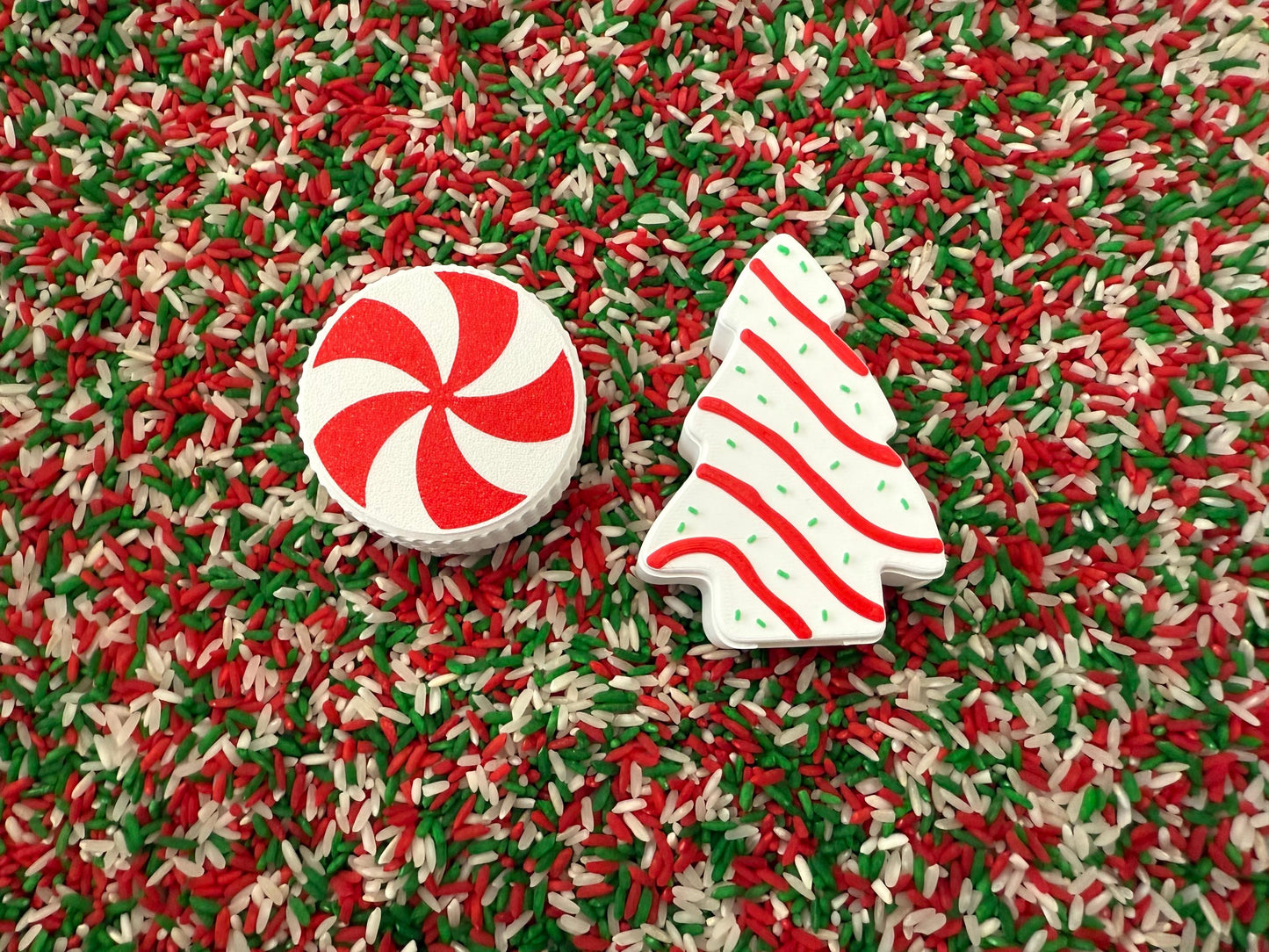 May include: A festive Christmas-themed sensory bin with a red and white peppermint candy and a white Christmas tree with red stripes. The background is a mix of red, green, and white rice.