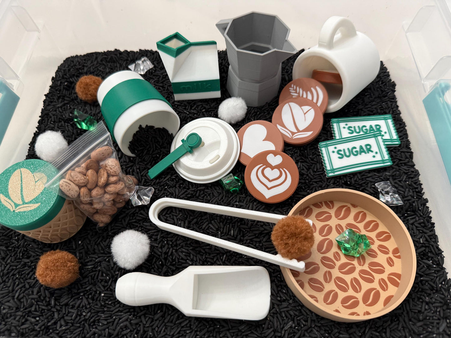 May include: A collection of coffee-themed toys, including a white mug, a gray moka pot, and a green and white coffee cup. There are also coffee beans, sugar packets, and latte art coasters. The items are arranged on a black surface.