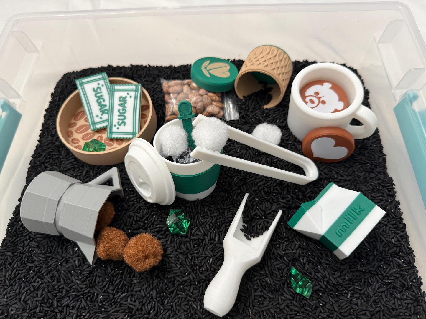 May include: A collection of toy coffee shop items, including a white mug with a bear design, a miniature coffee maker, and sugar packets. The items are arranged on a bed of black rice, with green and brown accents.