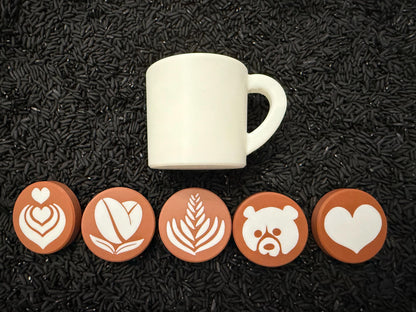 May include: A white ceramic mug with a curved handle sits above five round, brown coasters. Each coaster features a different white latte art design: a heart, coffee bean, fern, bear, and heart.
