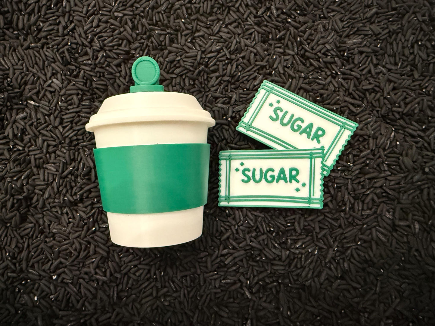 May include: A white and green coffee cup with a green sleeve and lid, next to two sugar packets. The sugar packets are white with green borders and the word "SUGAR" printed on them.