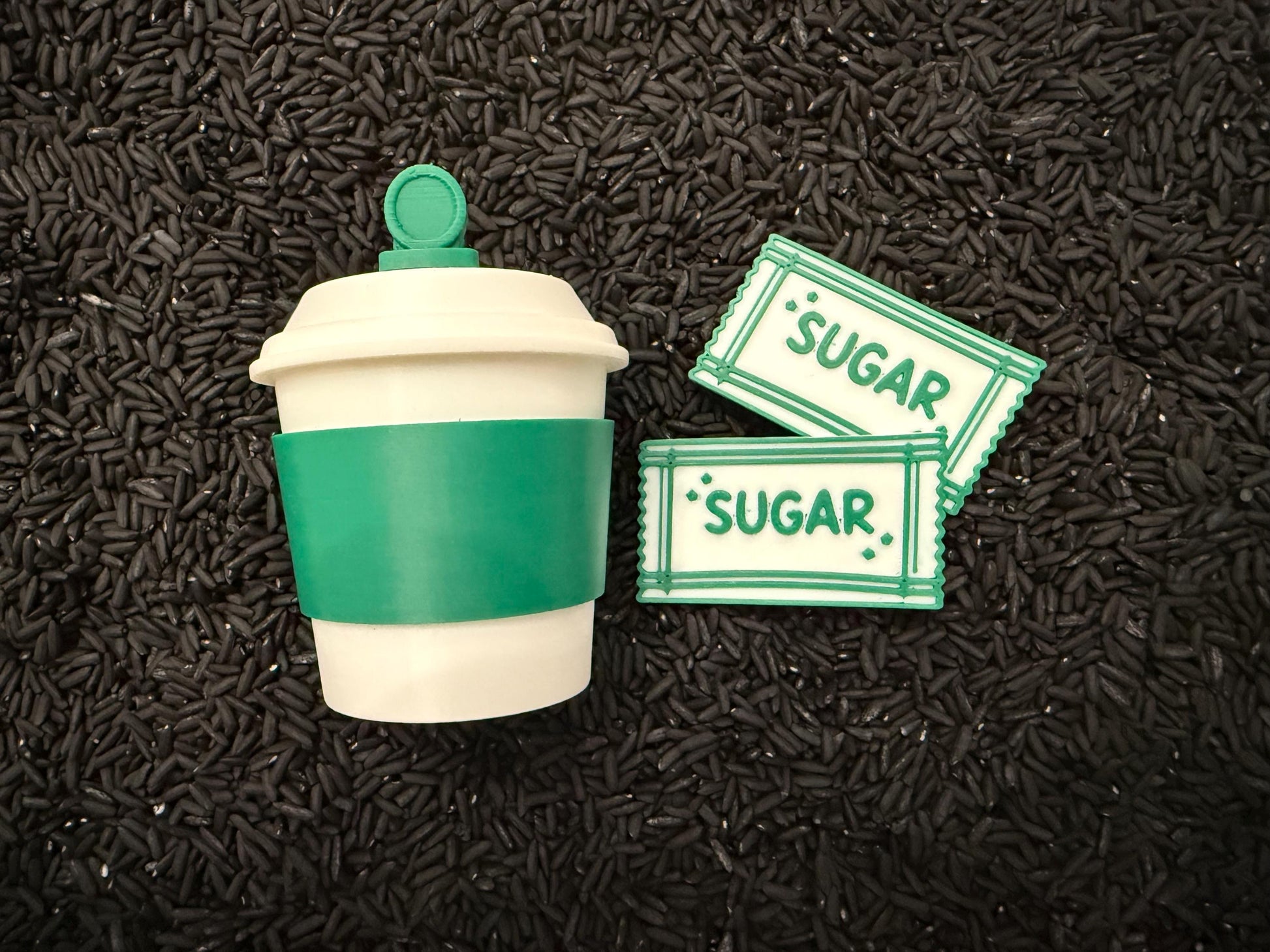 May include: A white and green coffee cup with a green sleeve and lid, next to two sugar packets. The sugar packets are white with green borders and the word "SUGAR" printed on them.