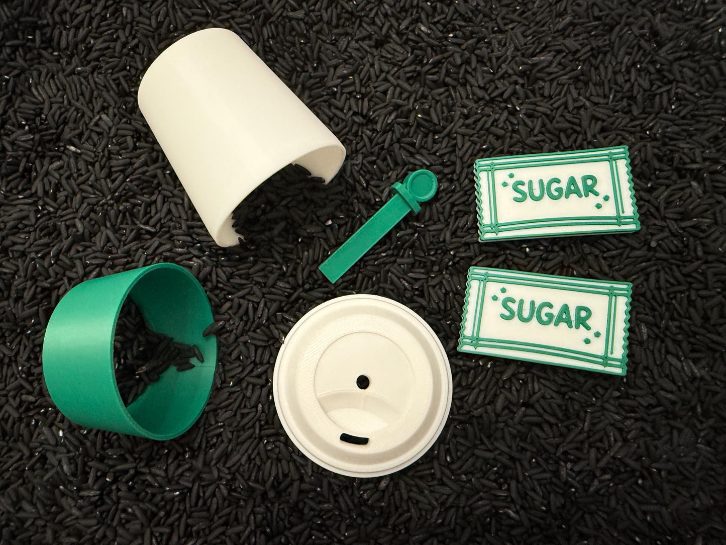 May include: A disassembled coffee cup set with a white cup, green sleeve, and lid. A green stirrer and two sugar packets with "SUGAR" text are also present. The items are on a black surface.