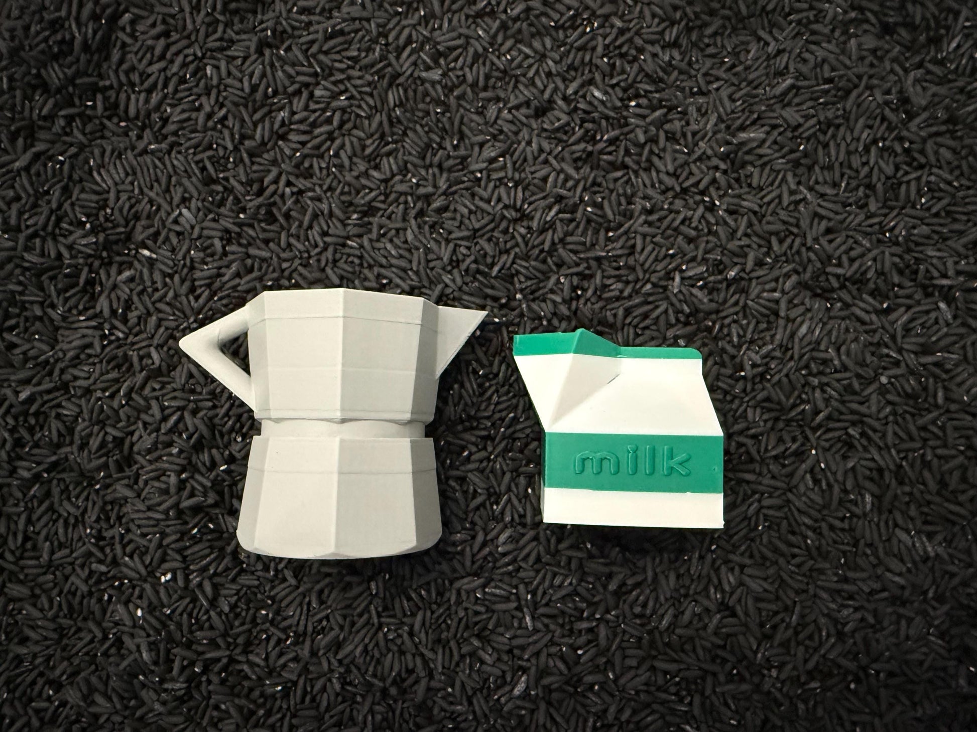 May include: A gray, geometric coffee maker and a white and green milk carton are displayed on a bed of black rice. The milk carton has the word "milk" printed on it. The objects are likely decorative.