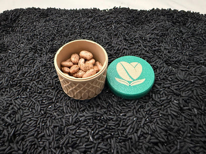 May include: A beige, patterned container filled with brown beans, next to a teal lid with a white coffee bean heart logo. The objects rest on a bed of black rice.