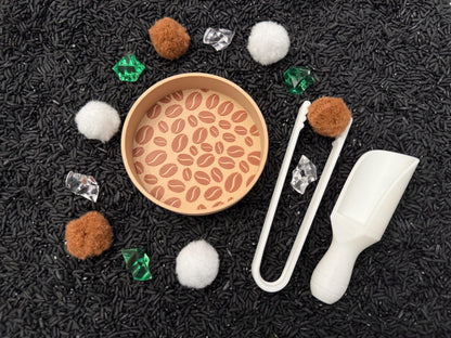 May include: A sensory play set with a round wooden tray featuring a coffee bean design. The set includes brown pom-poms, white tongs, a white scoop, clear and green gems, and white pom-poms, all arranged on a bed of black rice.