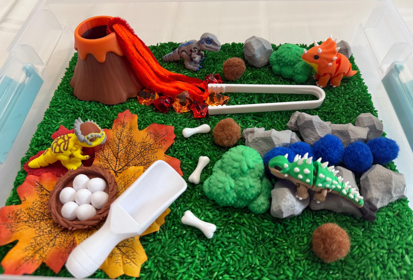 May include: A dinosaur-themed sensory bin with a volcano, dinosaurs, rocks, leaves, eggs, and bones. The volcano erupts with orange lava. The scene includes a variety of colorful toy dinosaurs and a white scoop.