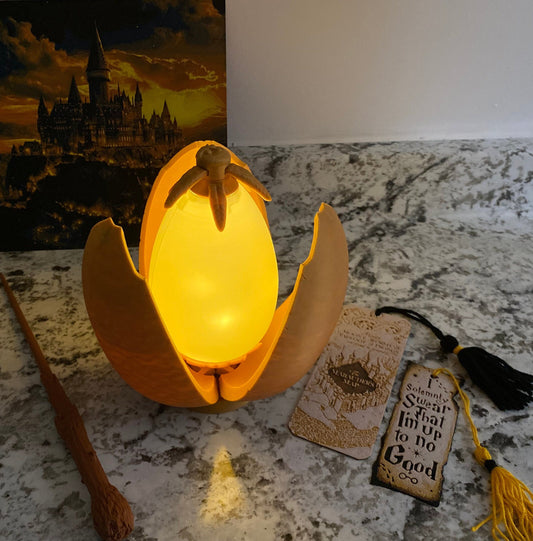May include: A golden egg-shaped lamp with a glowing light inside. The egg is open and the top half is resting on the bottom half. The lamp is on a table with a bookmark that says "I solemnly swear that I'm up to no good" and a wand.