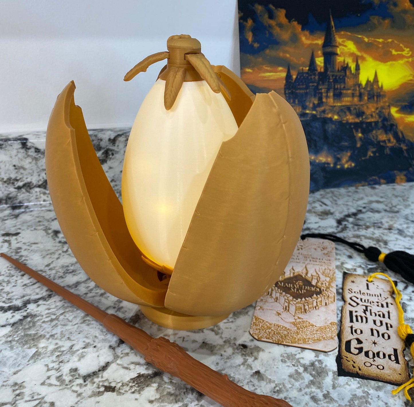May include: A golden egg-shaped lamp with a glowing white light inside. The egg is cracked open, revealing the light source. The lamp is sitting on a table with a brown wooden wand and a map of Hogwarts.