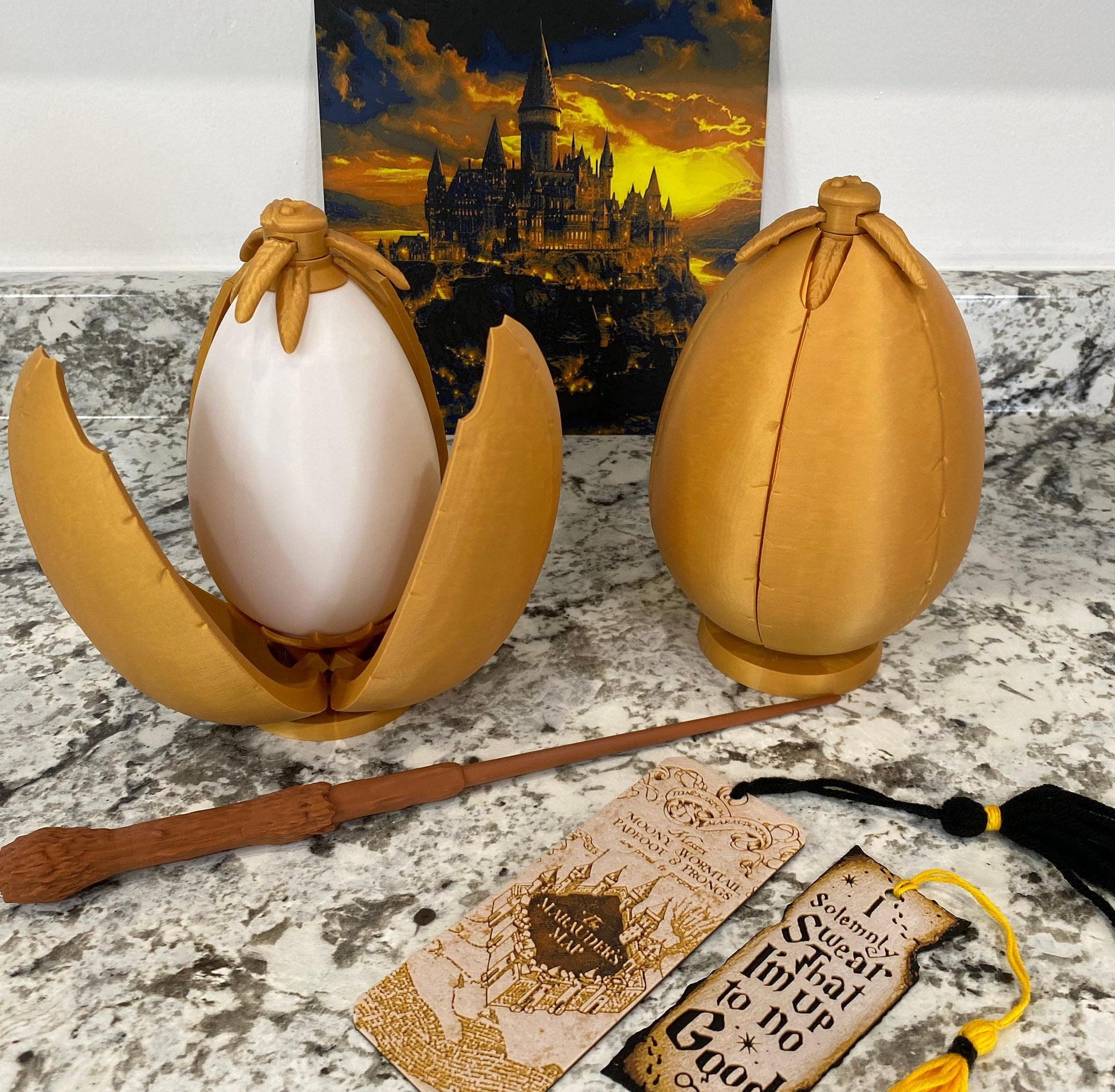 May include: Two golden egg-shaped containers, one open to reveal a white egg-shaped object inside. The containers are sitting on a light gray surface. A brown wooden wand and a bookmark with the text "Marauder's Map" are also visible.