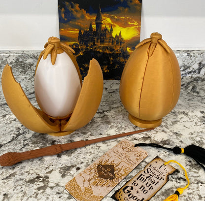May include: Two golden egg-shaped containers, one open to reveal a white egg-shaped object inside. The containers are sitting on a light gray surface. A brown wooden wand and a bookmark with the text "Marauder's Map" are also visible.