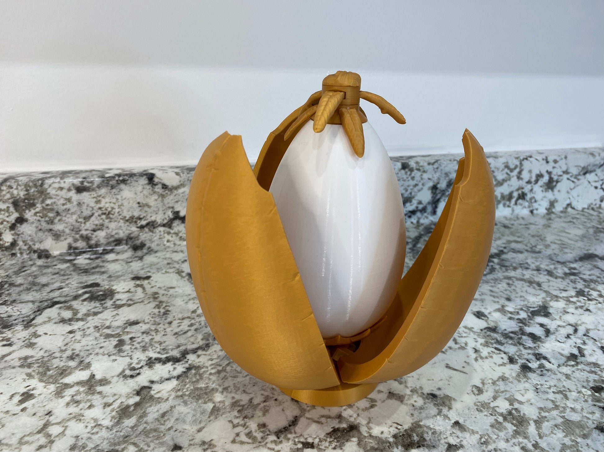 May include: A gold-colored plastic egg with a white egg inside. The egg is open and the white egg is visible. The egg is a toy.