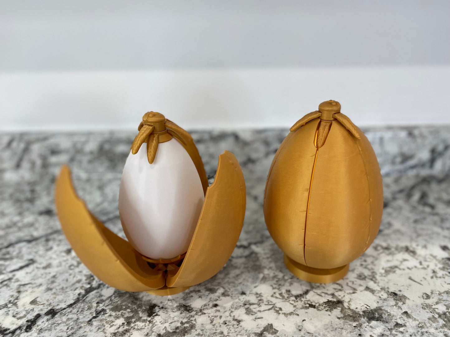 May include: Two gold-colored egg-shaped containers with a hinged opening. One container is open, revealing a white egg-shaped object inside.