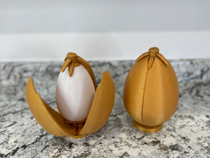 May include: Two gold-colored egg-shaped containers with a hinged opening. One container is open, revealing a white egg-shaped object inside.