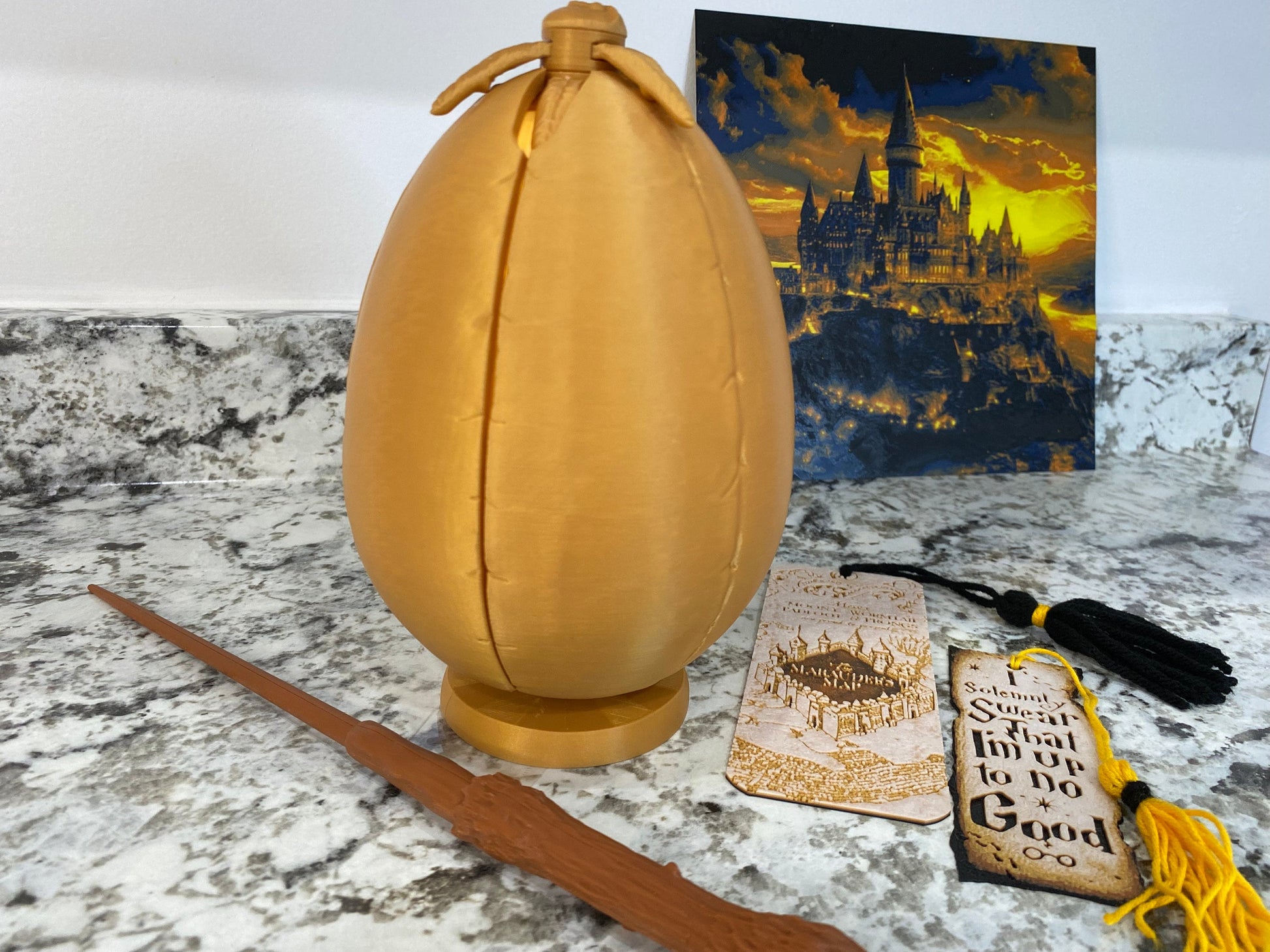 May include: A golden egg-shaped object with a small opening at the top. The egg is sitting on a table next to a wooden wand, a bookmark with a map of Hogwarts, and a bookmark with the text "It is solemn, sweet, that I'm up to no good".