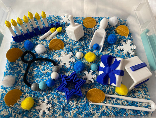May include: A sensory bin filled with blue and white rice, white snowflakes, blue and yellow pom poms, and other small toys. The toys include a blue and white dreidel, a blue gift box with a white lid, a blue star-shaped cookie cutter, a white scoop, and a blue and white menorah with yellow candles.