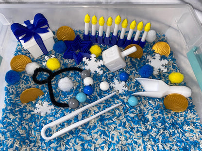 May include: A blue and white sensory bin filled with rice, blue and white pom poms, gold coins, and a variety of blue and white toys, including a dreidel, a menorah, a star of David, and a gift box.