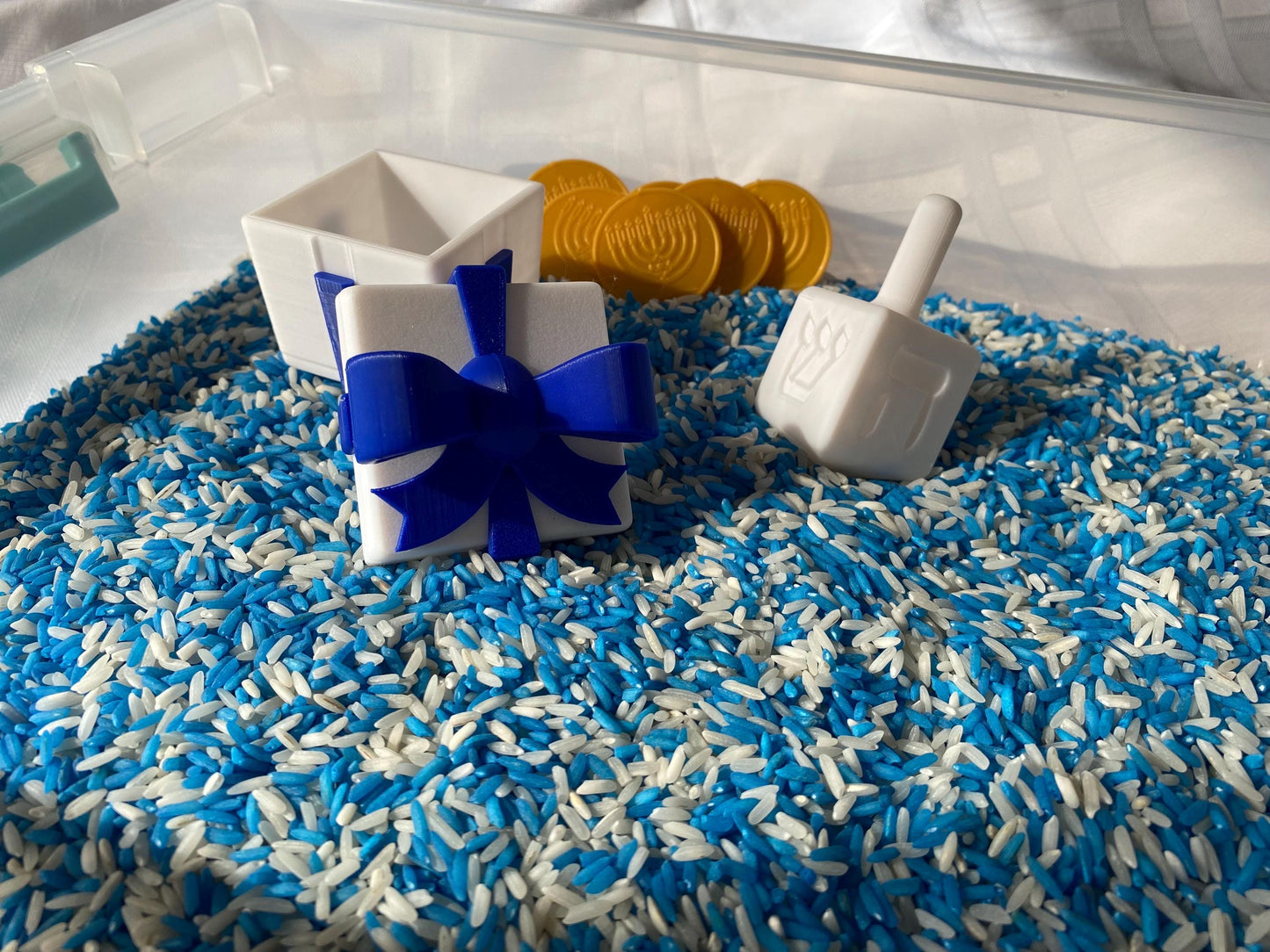 May include: A white plastic gift box with a blue bow sits in a clear container filled with blue and white rice. A white plastic dreidel and gold coins are also in the container.
