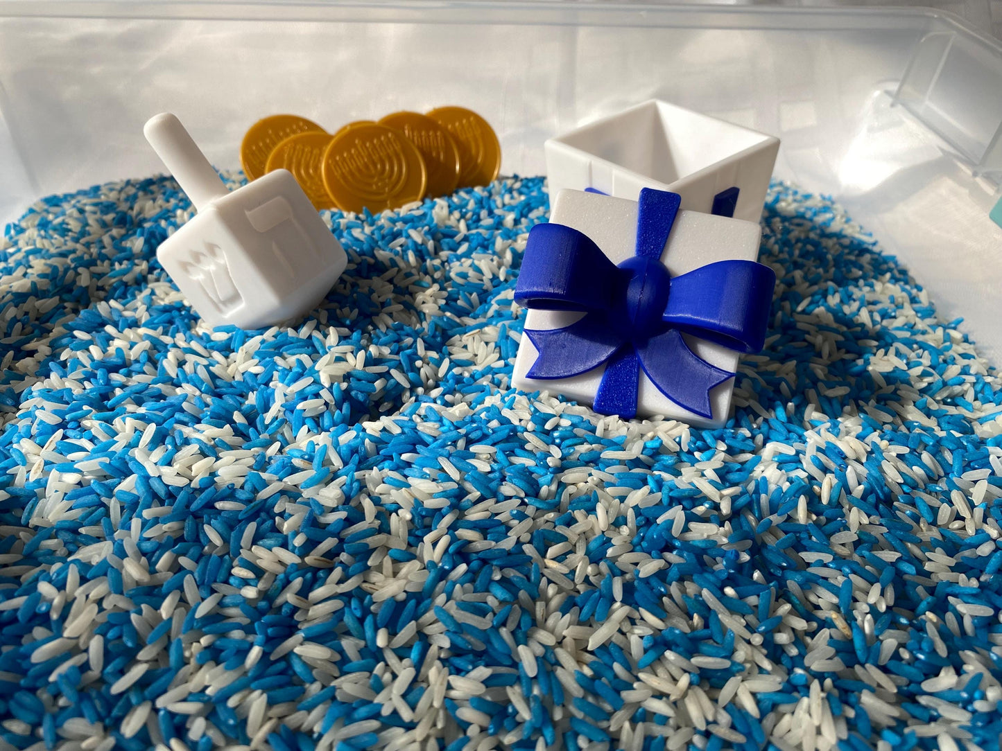May include: A white plastic dreidel, a white gift box with a blue bow, and gold coins with a menorah design are partially submerged in a blue and white rice mixture.