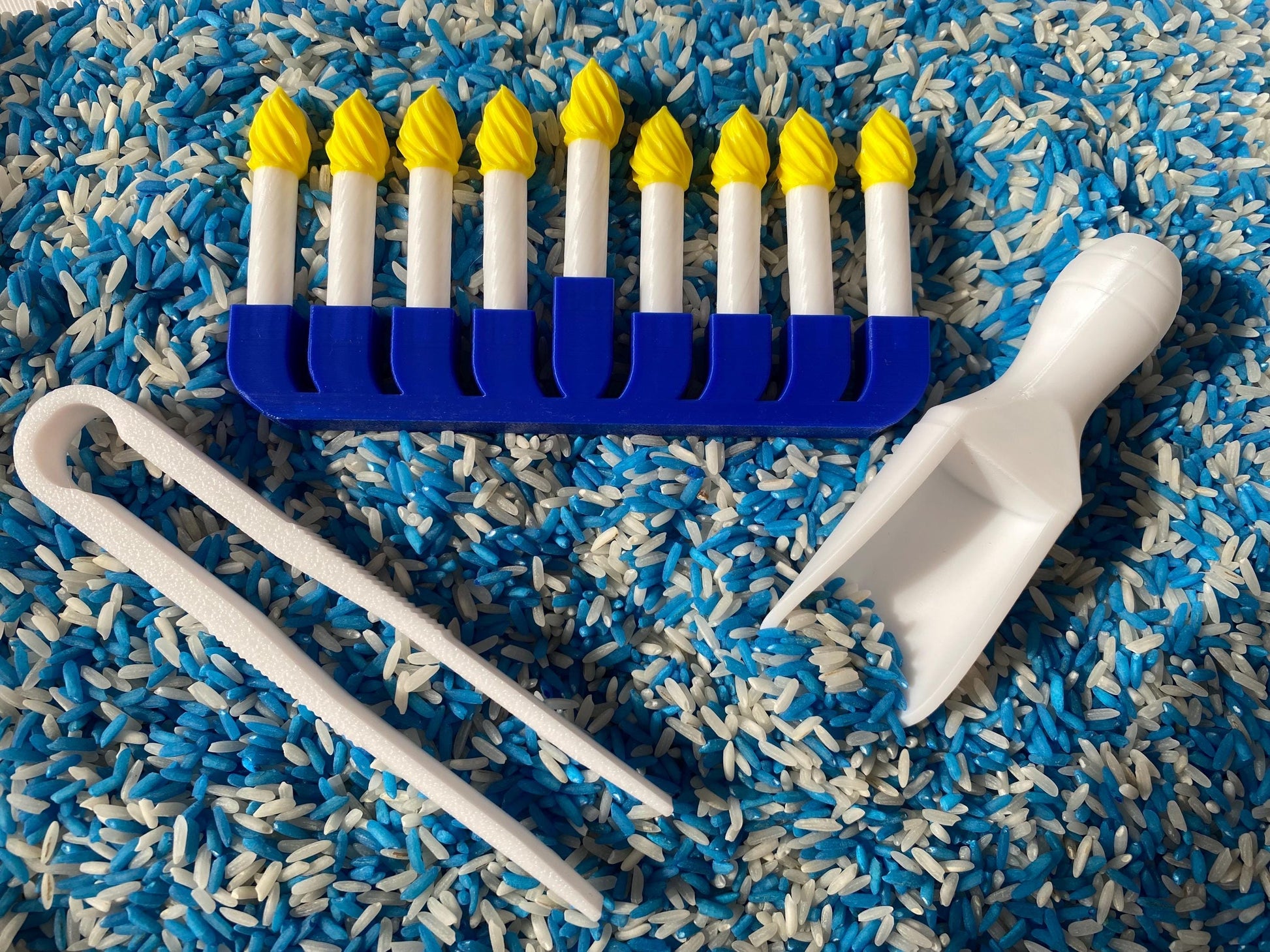 May include: A blue menorah with nine white candles and yellow flames sits on a bed of blue and white rice. A white scoop and tongs are also included.