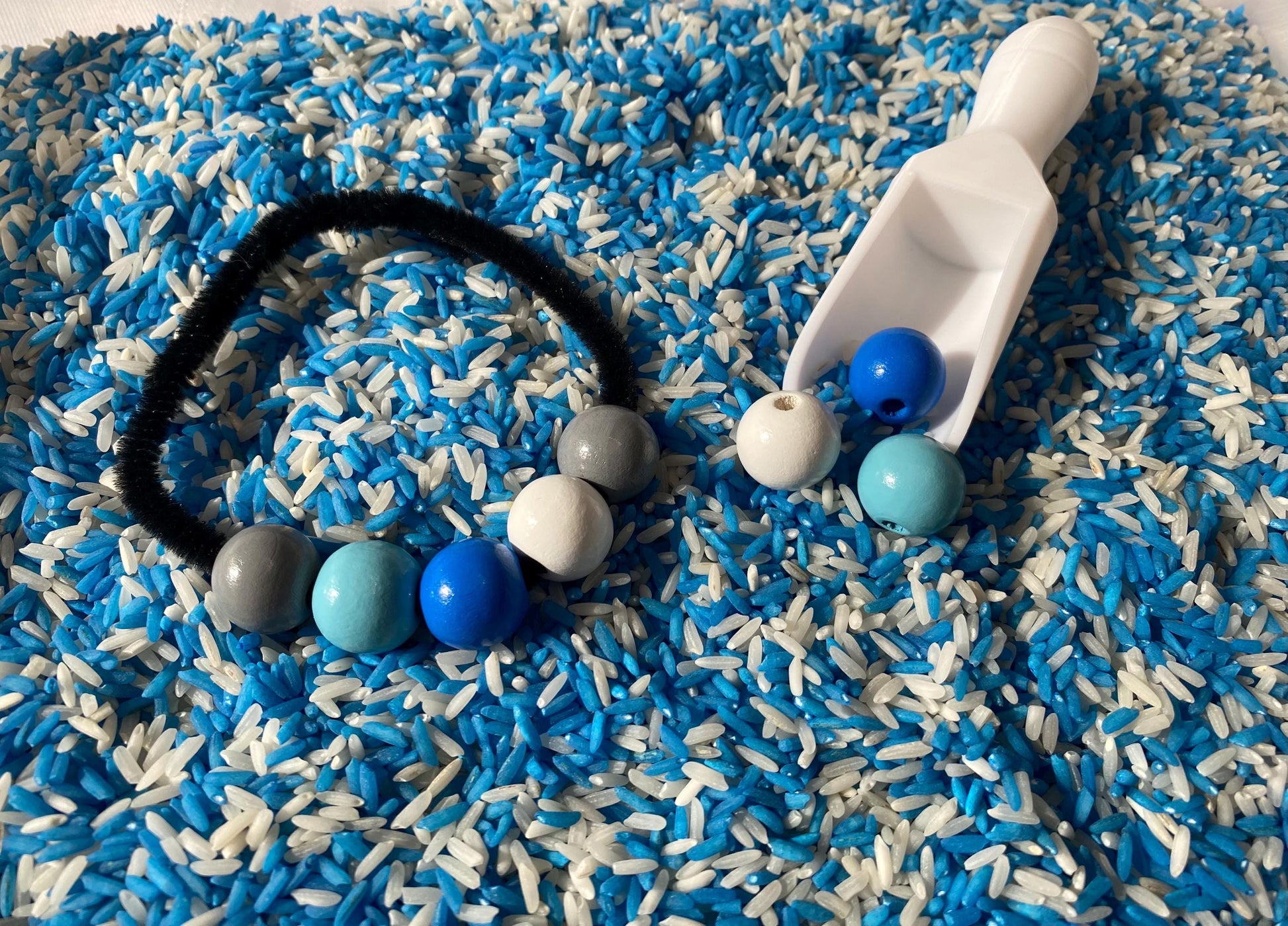 May include: A sensory bin filled with blue and white rice. A black pipe cleaner with gray, blue, and white beads is on top of the rice. A white plastic scoop with blue and white beads is in the bin.