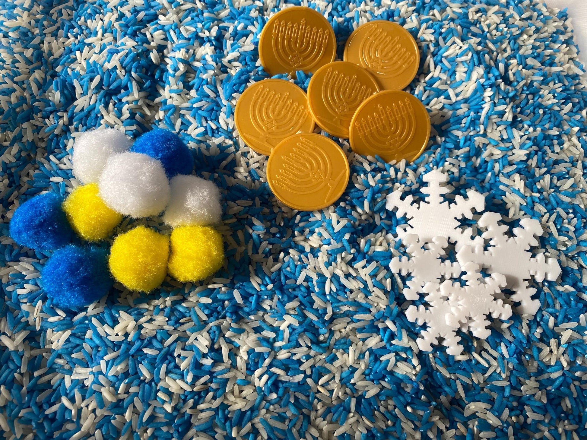 May include: A sensory bin filled with blue and white rice, featuring yellow and blue pom-poms, white snowflakes, and gold coins with a menorah design.