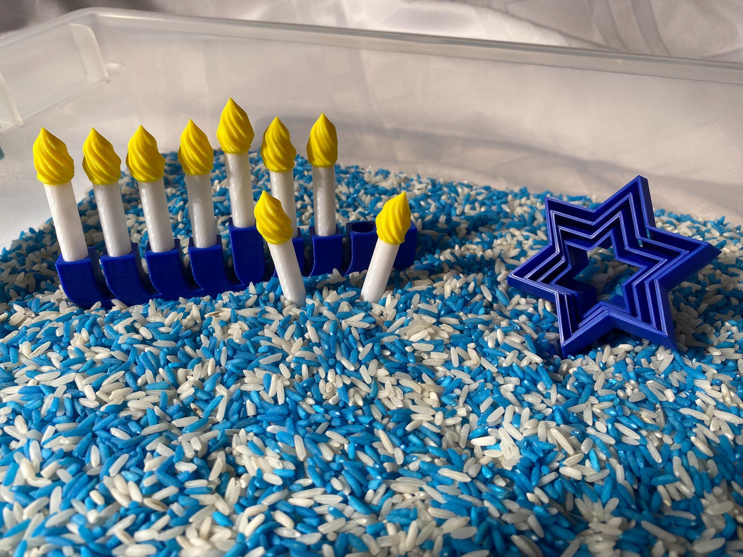 May include: A blue menorah with yellow candles sits in a sensory bin filled with blue and white rice. A blue star of David cookie cutter is also in the bin.