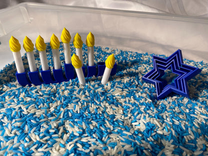 May include: A blue menorah with yellow candles sits in a sensory bin filled with blue and white rice. A blue star of David cookie cutter is also in the bin.